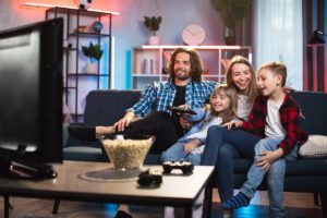 Smiling young parents with two kids sitting on couch and watching TV. Happy caucasian family in casual wear spending evening time together at home.