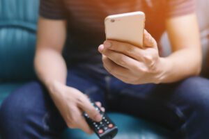 A man looking at his mobile phone, holding a remote in his other hand.